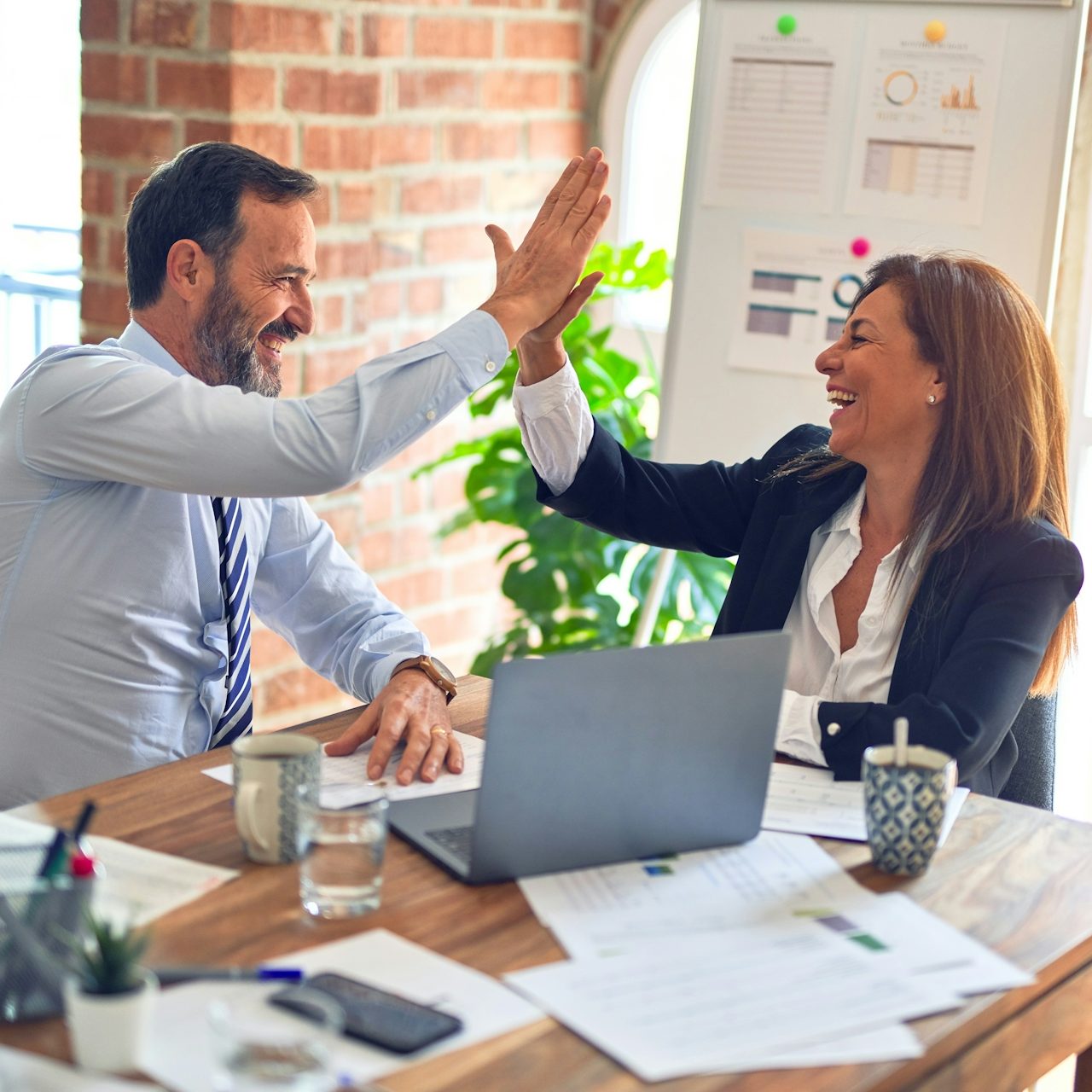 Zwei Personen lachen und gratulieren sich in einem modernen Büro zu einer erfolgten Kooperation. Unterlagen liegen auf dem Tisch.