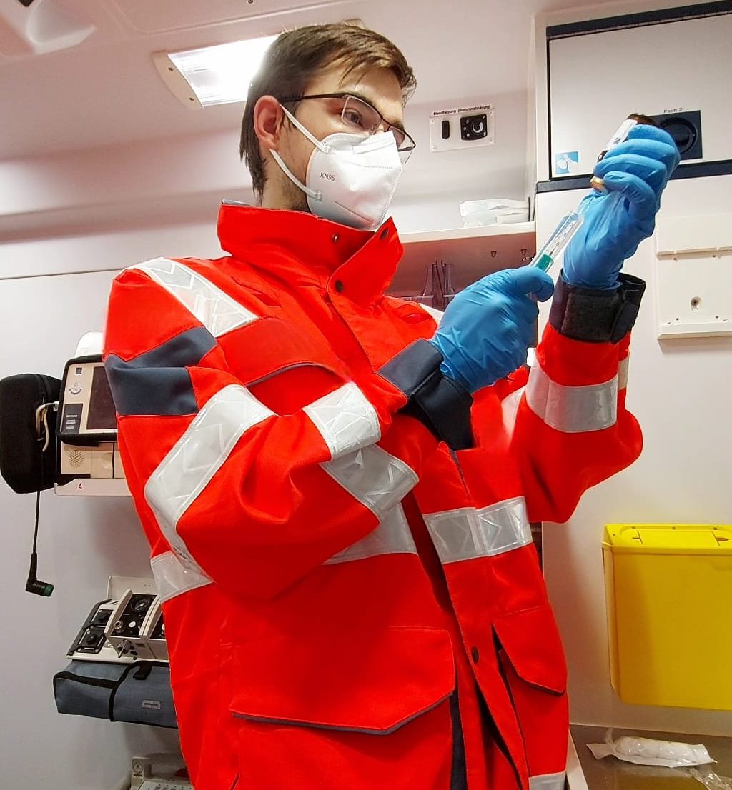 Ein Sanitäter steht in einem Rettungswagen. An der Wand befinden sich medizinische Geräte. Ein Medikament wird in einer Spritze aufgezogen.