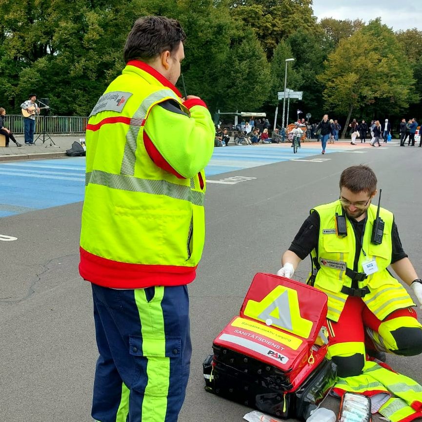 Zwei Sanitäter*innen sind in Leipzig unterwegs und versorgen eine hilfsbedürftige Person am Straßenrand.