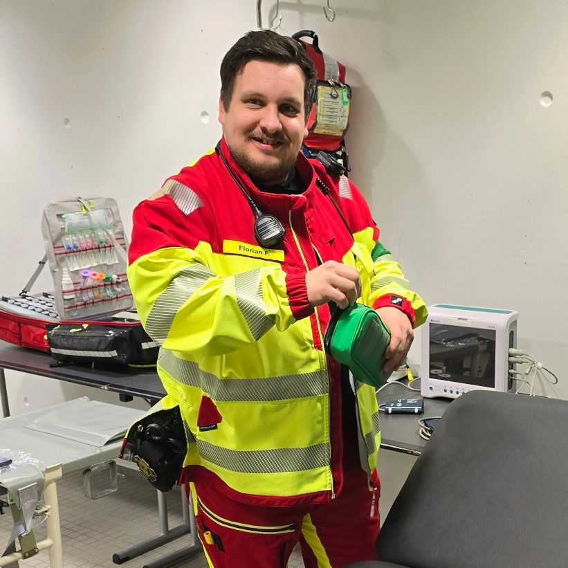 Ein Sanitäter in Einsatzkleidung steht in einem Sanitätsraum mit Notfallrucksack und EKG-Gerät. Er bereitet den Einsatz vor.