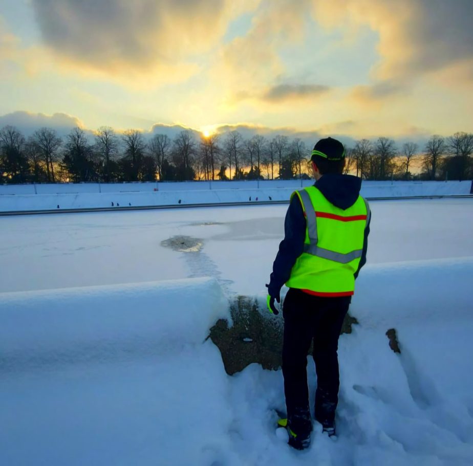 Person in Warnweste steht an gefrorenem Gewässer bei Sonnenuntergang. Schnee umgeben.