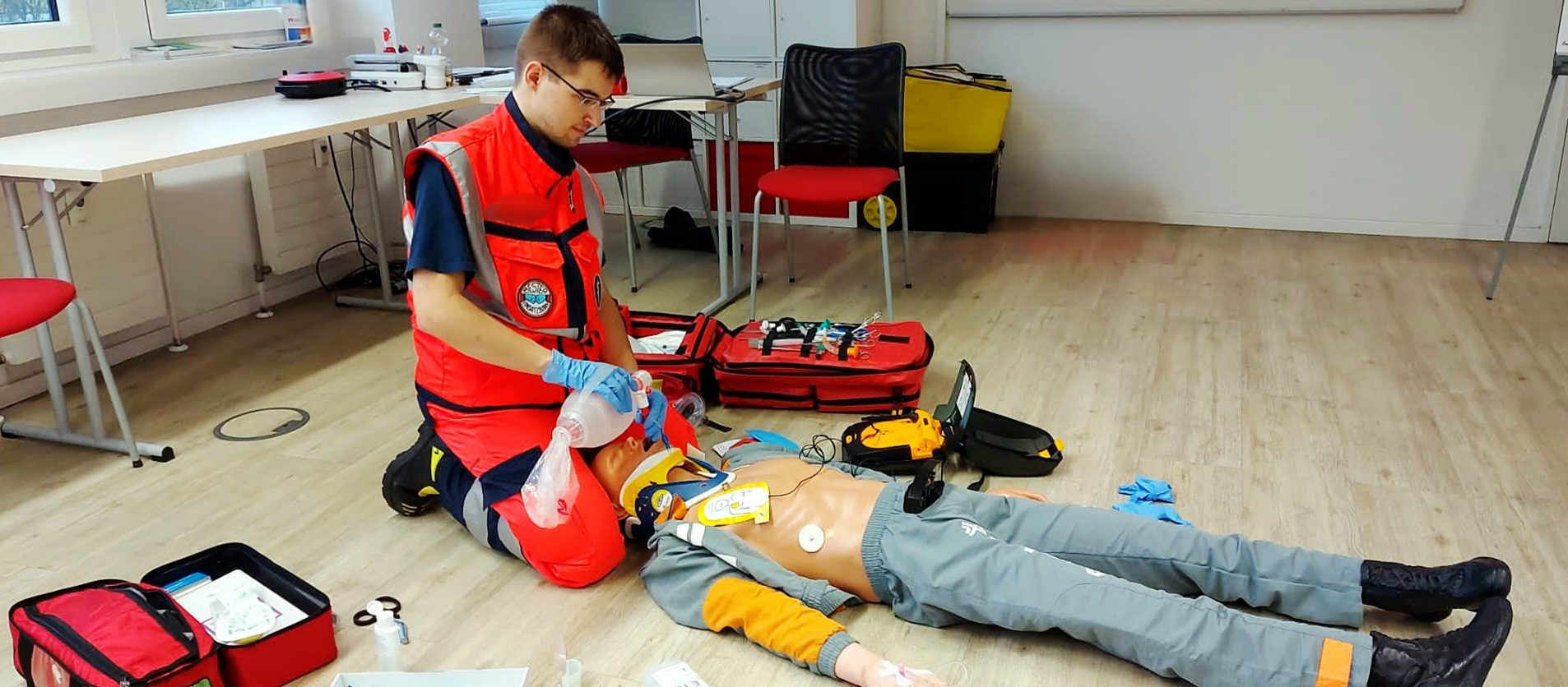 Ein Sanitäter führt Erste Hilfe bei einem simulierten Patienten durch. Ein AED ist angeschlossen. Der Patient wird beatmet.