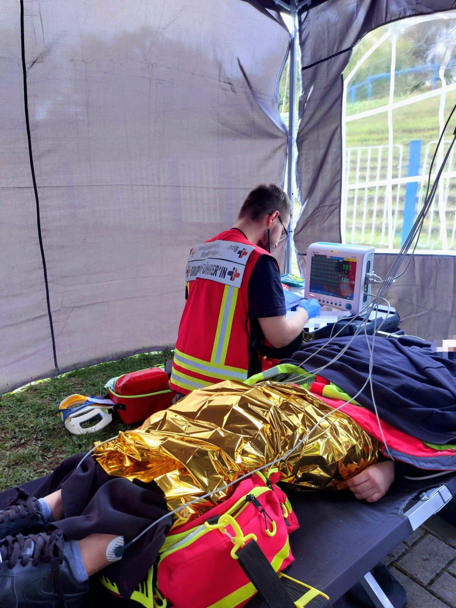 Ein Sanitäter mit roter Weste (Gruppenführer*in) versorgt eine*n liegende*n Patient*in und wertet ein EKG aus.