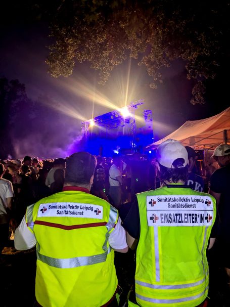 Zwei Sanitäter stehen vor einem festlichen Ereignis mit bunten Lichtern und Menschenmenge. Im Hintergrund ist eine Bühne zu sehen.