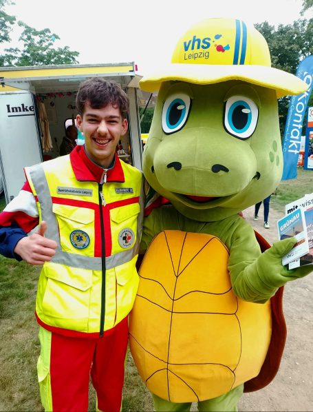 Ein Sanitäter des SKL posiert lächelnd mit einem Maskottchen der VHS in einem Schildkrötenkostüm.