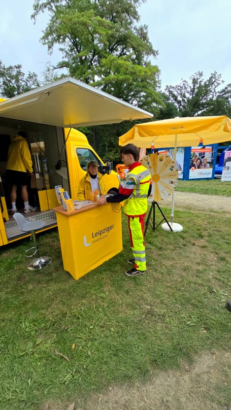 Ein Sanitäter spricht mit einer Person an einem Infostand der Leipziger Stadtwerke am Rand des Parkfests.