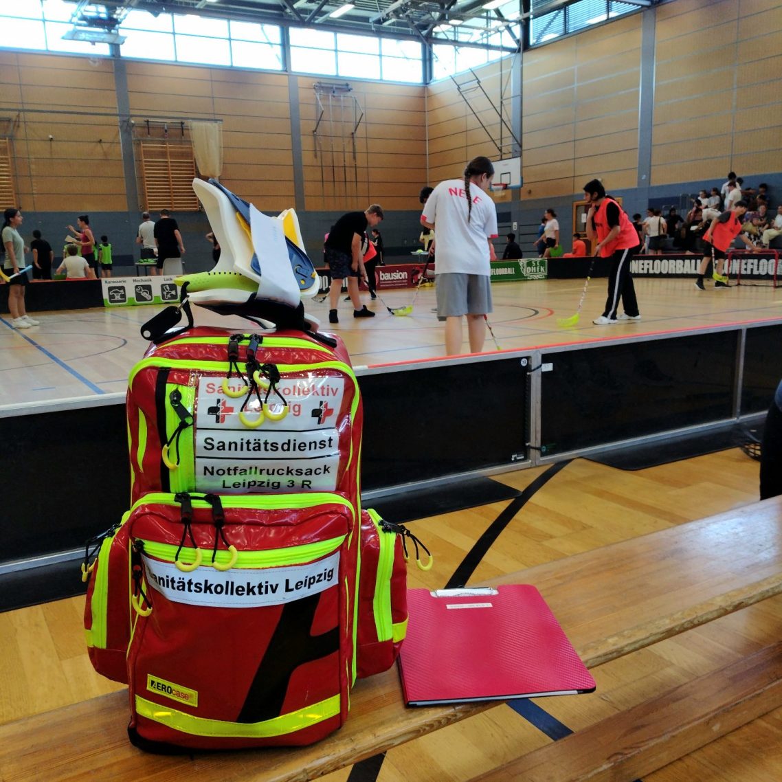 Auf einer Bank steht ein Notfallrucksack bereit für die Erstversorgung von Sportler*innen. Im Hintergrund wird Floorball gespielt.