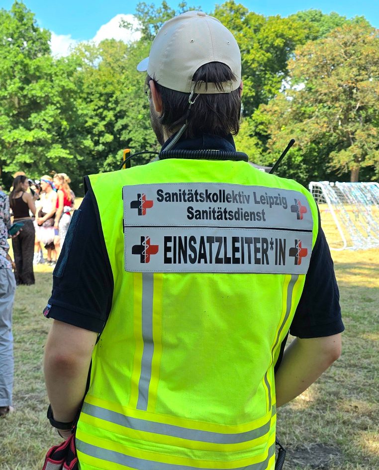 Ein Sanitäter des SKL steht in einem Park und beobachtet eine Sportveranstaltung. Er trägt eine gelbe Weste mit Aufschrift Einsatzleiter*in.