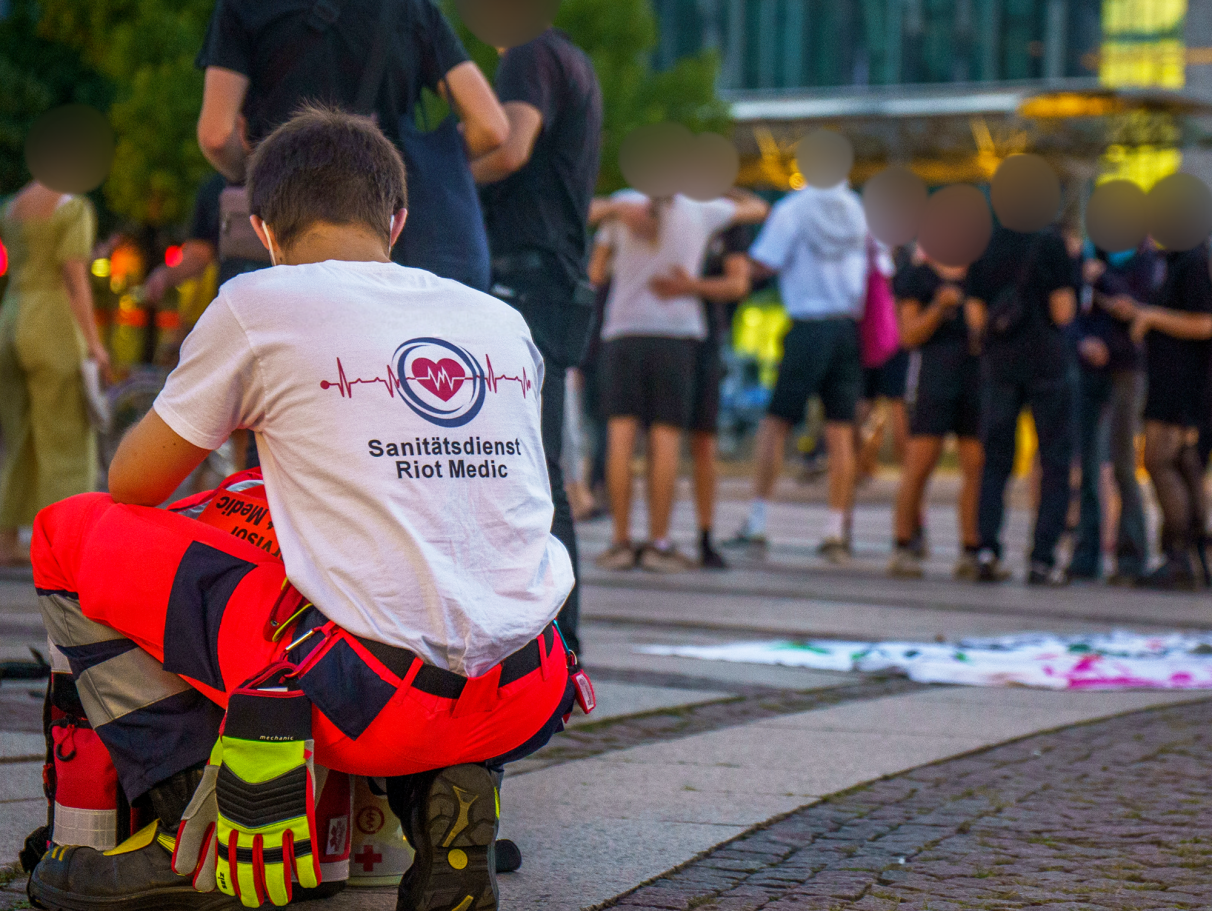 Junger Sanitäter in roter Hose, kniend, beobachtet eine Menschenmenge im Hintergrund.