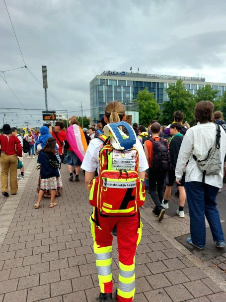 Einsatzkraft des Sanitätskollektivs mit Notfallrucksack geht neben einer Demo mit trans* und FLINTA*-Flaggen mit. Sie sichert die Demo ab.