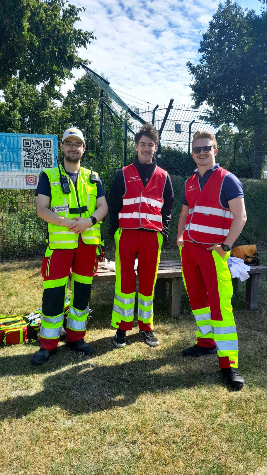 Drei Sanitäter*innen stehen auf einem Sportplatz und sichern eine Schulsportveranstaltung ab. Sie lächeln in die Kamera.
