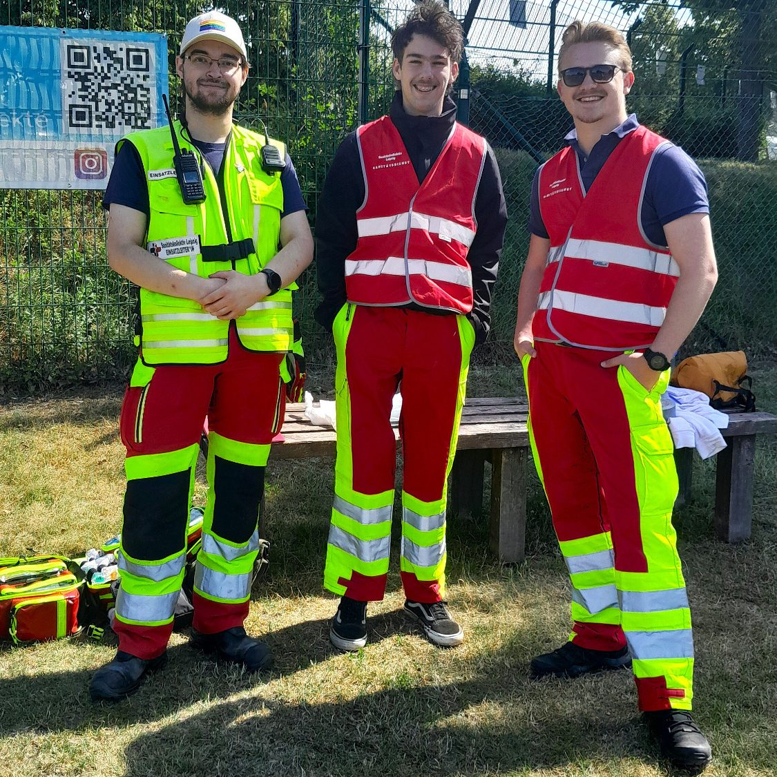 3 Sanitäter*innen sichern eine Sportveranstaltung ab. Sie stehen auf dem Sportplatz und haben einen Notfallrucksack dabei.