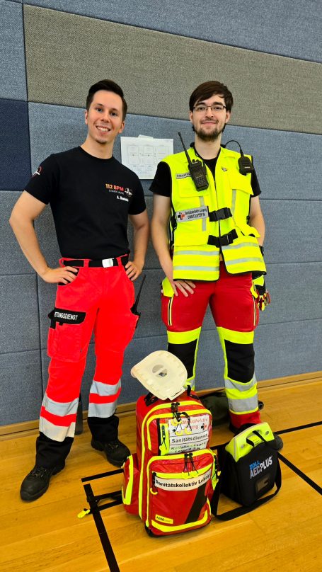 Zwei Sanitäter*innen mit Ausrüstung stehen in einer Sporthalle und lächeln in die Kamera. Sie sind auf einem Sanitätsdienst.