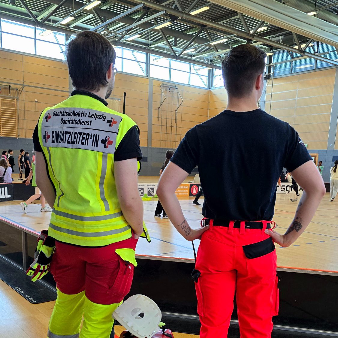 2 Sanitäter*innen stehen in Dienstkleidung am Rande des Spielfeldes und beobachten das Turnier in Basketball und Floorball.