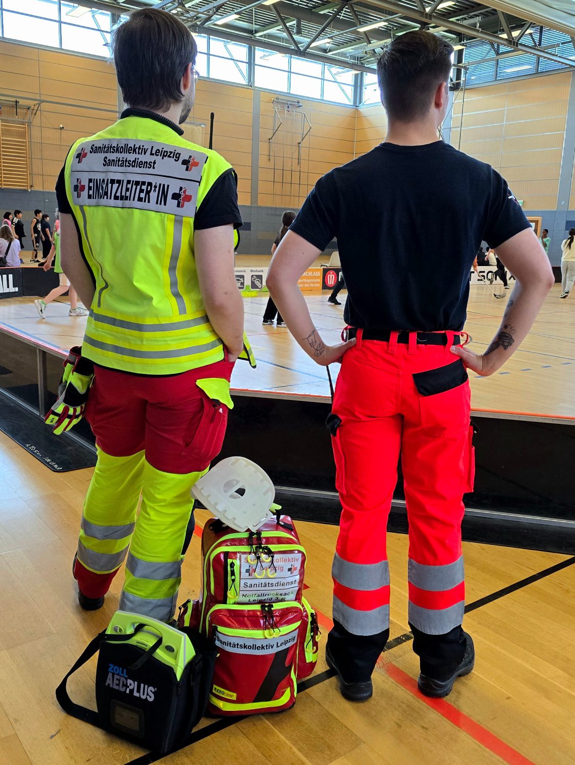Zwei Rettungskräfte in leuchtenden Uniformen beobachten ein Sportereignis in einer Sporthalle. Sie haben einen Notfallrucksack dabei.