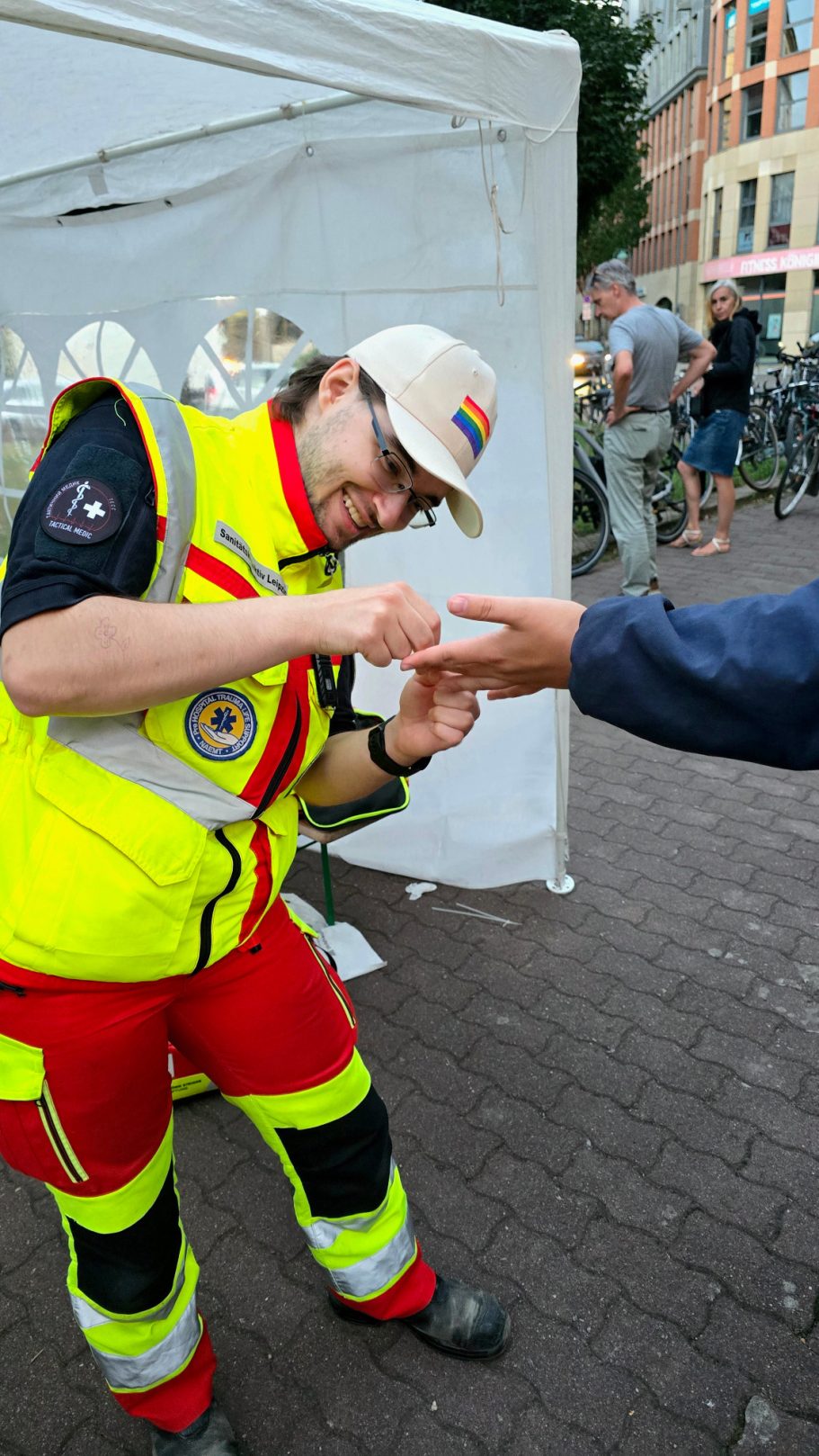 Ein Sanitäter in rot-gelber Einsatzkleidung steht vor einem Zelt bei eine Straßenfest und versorgt eine kleine Wunde an einem Finger.