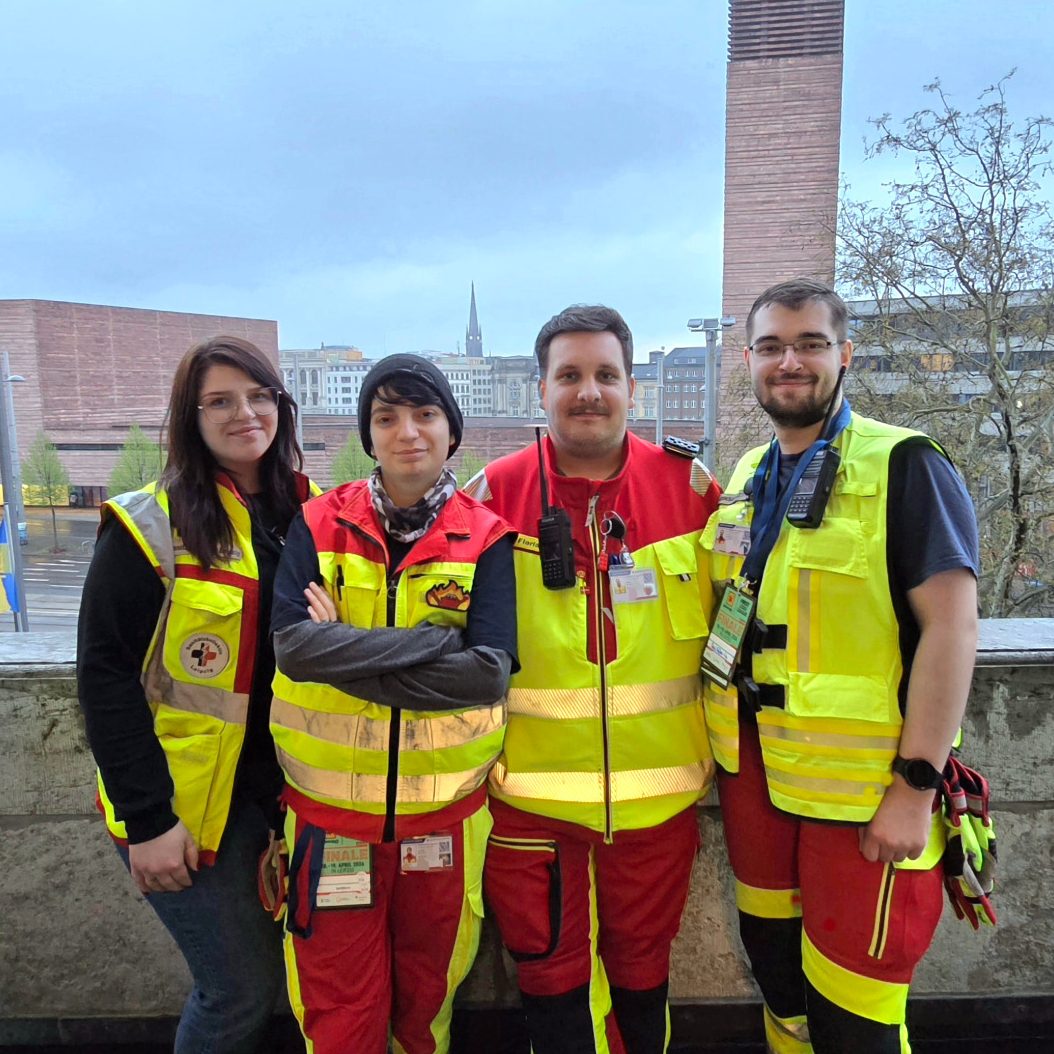4 Helfer*innen des Sanitätskollektiv Leipzig in rot-gelber Einsatzkleidung stehen auf dem Balkon des Neuen Rathaus Leipzig für ein Teamfoto.