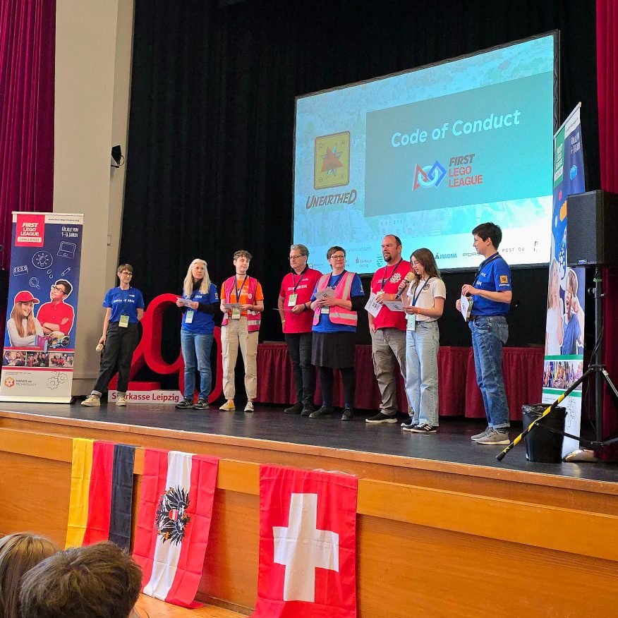 Eine Bühne mit den Flaggen von Deutschland, Österreich und Schweiz ist zu sehen. Das Organisationsteam der FIRST LEGO League spricht.