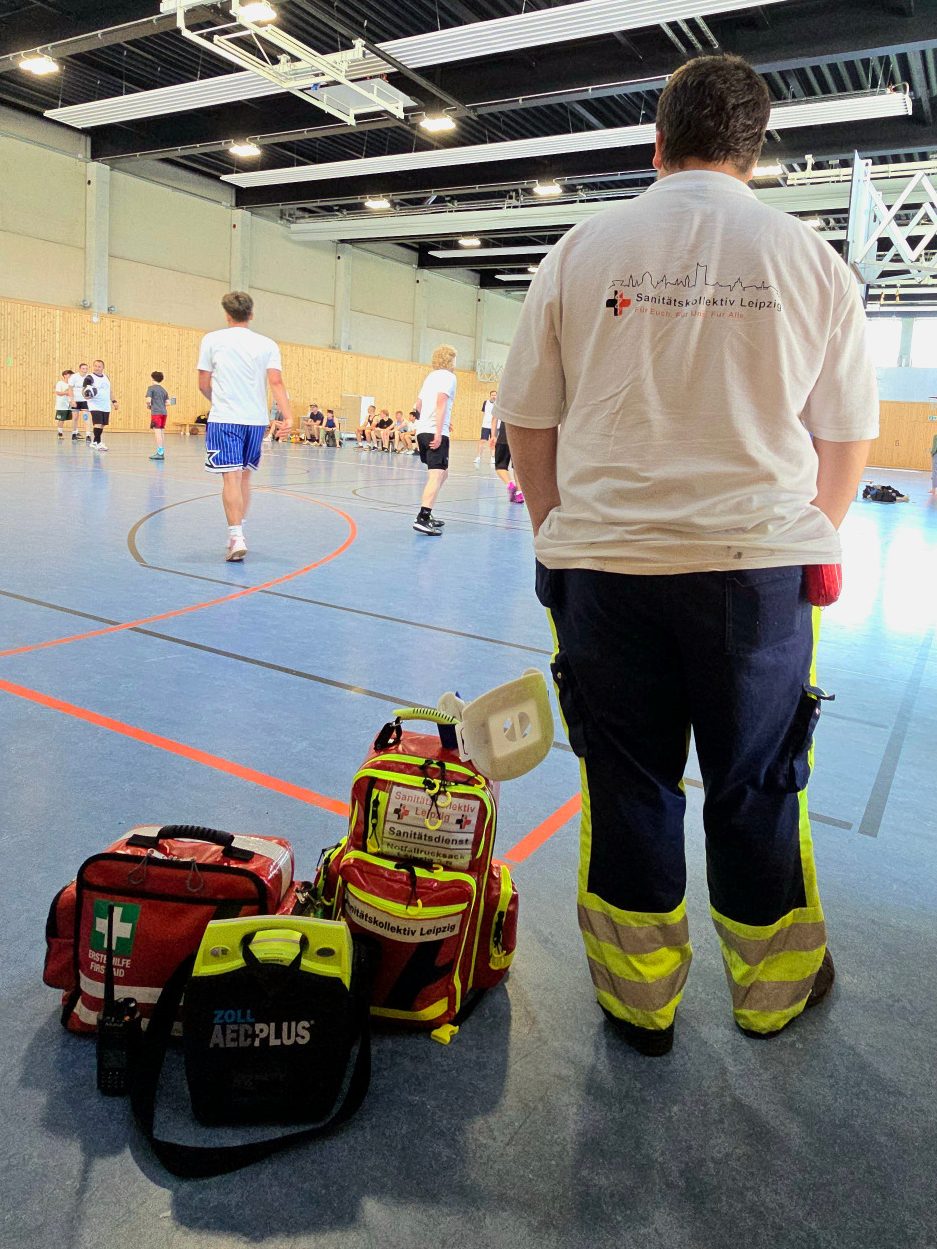 Sanitäter in Sporthalle mit medizinischer Ausrüstung (AED, Rucksäcke) im Vordergrund. Im Hintergrund sind Sportler beim Spielen zu sehen.