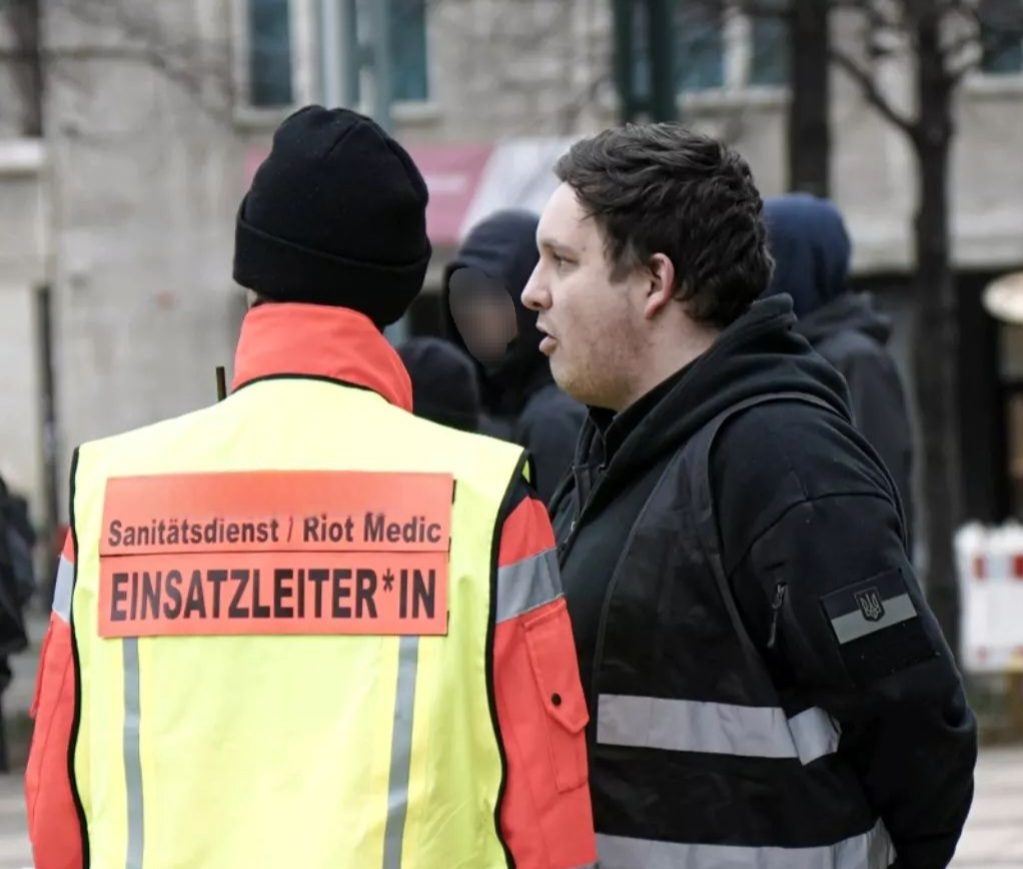 Ein Sicherheitschef einer Versammlung mit schwarzer Warnweste bespricht sich mit dem Einsatzleiter des Sanitätsdienstes vor Beginn der Demo.