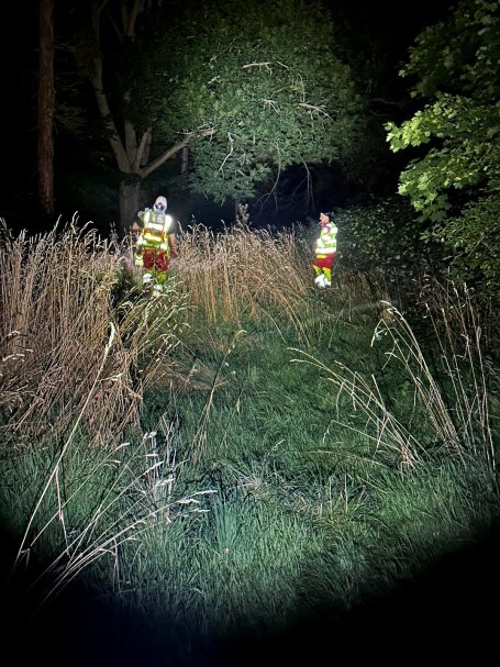 Zwei Sanitäter durchsuchen nachts einen Bereich im Park mit Taschenlampen. Sie suchen eine hilfelose Person.