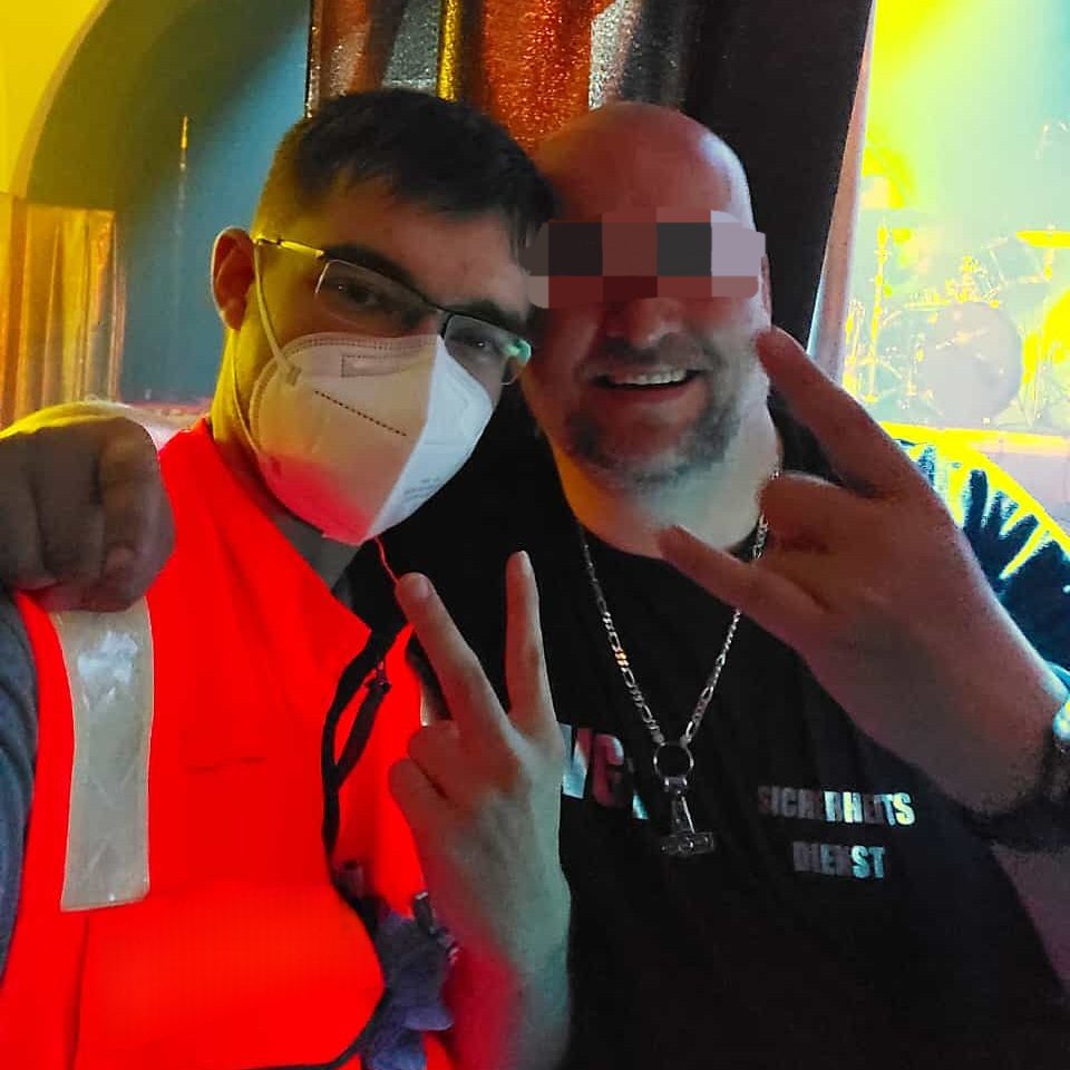 Ein Sanitäter in roter Einsatzkleidung und ein Mitarbeiter der Security stehen neben einer Bühne beim WGT und machen ein Selfi.