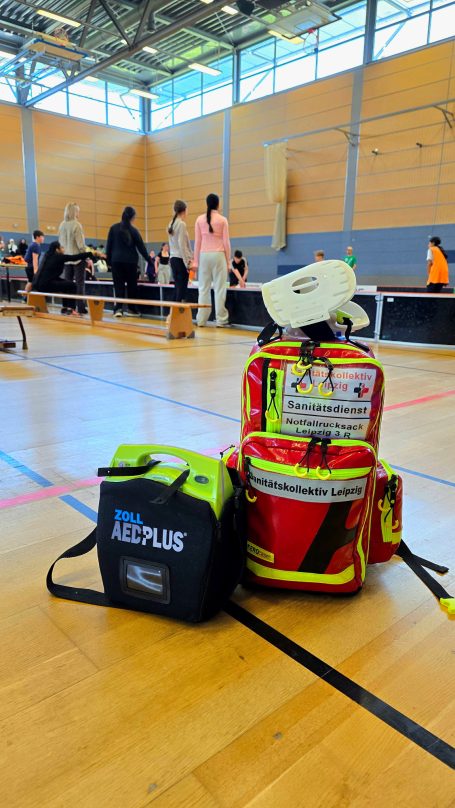 Rettungstasche und Defibrillator im Vordergrund, Menschen im Hintergrund in einer Sporthalle. Sie spielen Floorball.