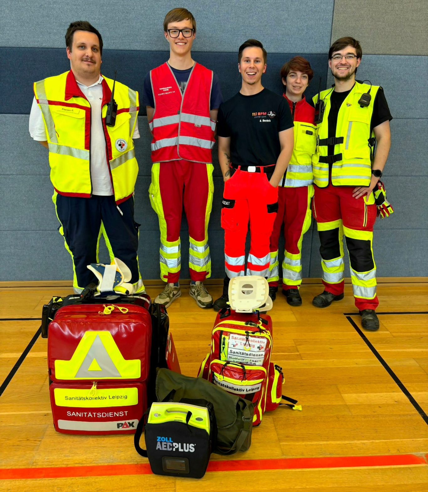 Gruppe von 5 Sanitäter*innen in Rettungsdienstkleidung steht in einer Sporthalle. Vor ihnen befinden sich mehrere Notfallrucksäcke und AED.